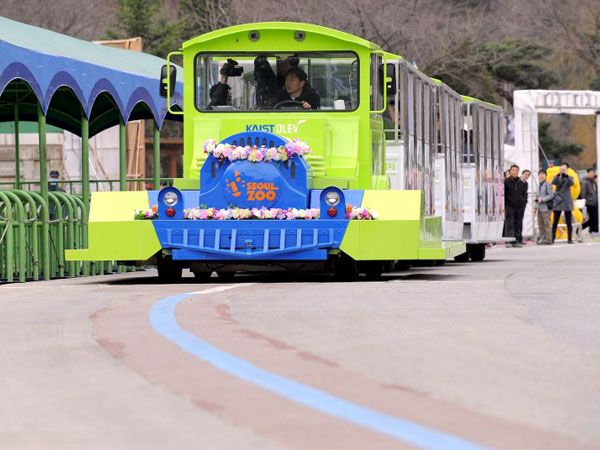 The Online Electric Vehicle (OLEV), towing three carriages, runs along a blue line under which power strips are buried for recharging, at an amusement