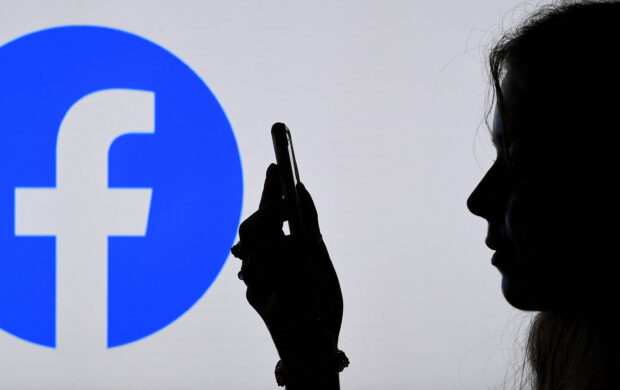 A woman browses her smartphone with Facebook logo at the background.